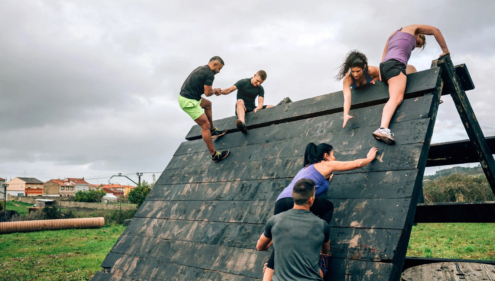 Gruppe von Hindernislauf-Teilnehmern hilft sich gegenseitig eine schräge Holzwand hinauf, Teamwork beim OCR Training.